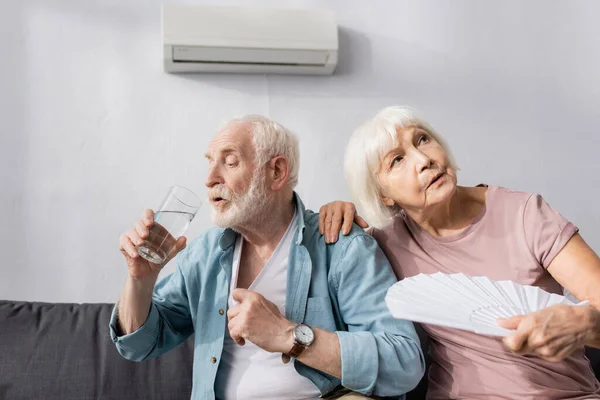 Femme âgée tenant ventilateur près du mari boire de l'eau sur le canapé à la maison — Photo de stock