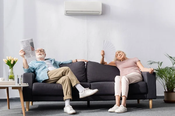 Elderly couple sitting on couch with fan and newspaper while feeling hot — Stock Photo