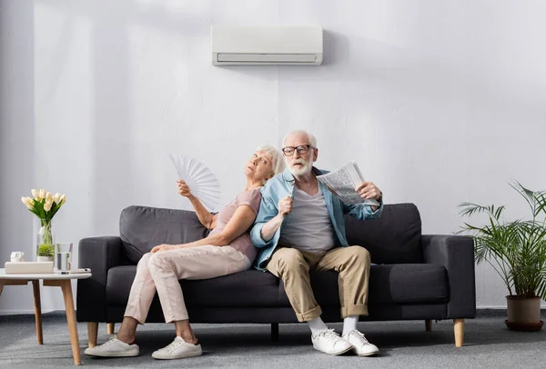 Mujer mayor sosteniendo ventilador mientras sufre de calor cerca de marido con periódico - foto de stock