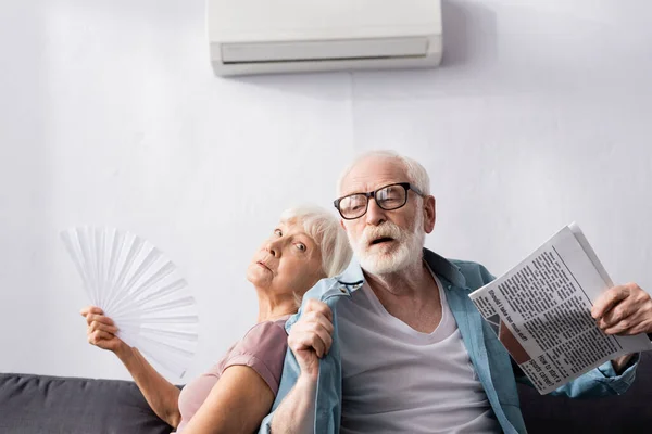 Hombre mayor sosteniendo periódico cerca de esposa cansada con ventilador bajo el aire acondicionado en la sala de estar - foto de stock