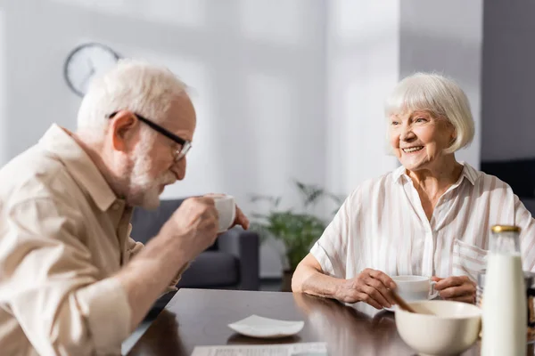 Выборочный фокус улыбающейся пожилой женщины, смотрящей на мужа, пьющего кофе дома — стоковое фото
