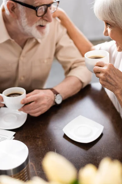 Foco seletivo de sorrir mulher sênior segurando xícara de café e abraçando o marido na cozinha — Fotografia de Stock