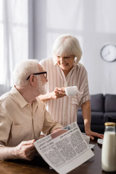 Selective focus of smiling senior woman holding cup of coffee near husband with newspaper — Stock Photo