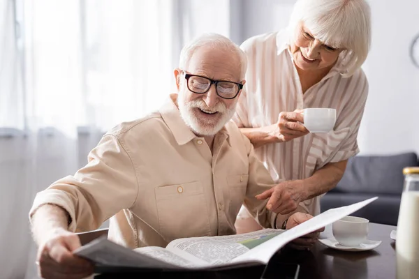 Selective focus of smiling senior man reading newspaper near wife with cup of coffee in kitchen — Stock Photo