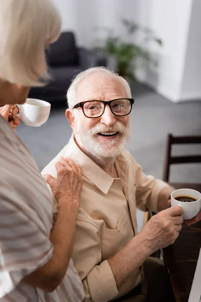 Foco seletivo do homem sênior segurando xícara de café perto da esposa em casa — Fotografia de Stock