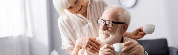 Panoramic shot of smiling senior woman embracing husband with cup of coffee at home — Stock Photo