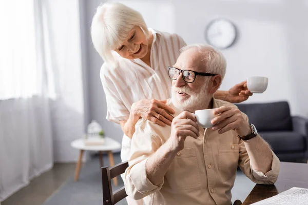 Lächelnde Seniorin hält Tasse Kaffee in der Hand und umarmt Ehemann beim Frühstück zu Hause — Stockfoto