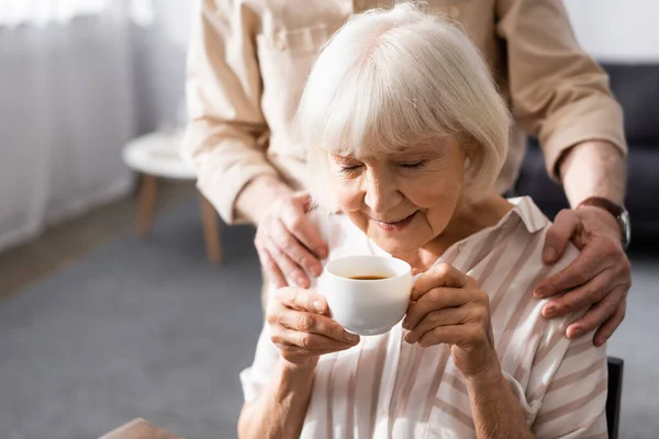 Выборочный фокус пожилого мужчины, обнимающего жену с чашкой кофе дома — стоковое фото