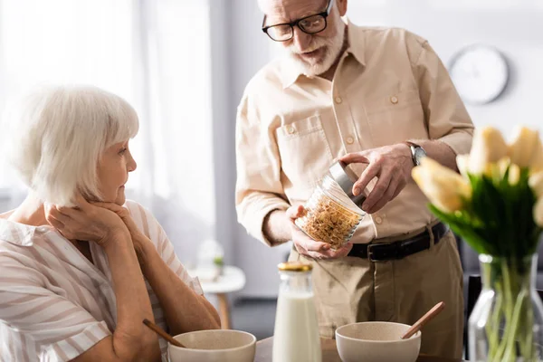 Foco seletivo do homem sênior frasco de abertura com cereais perto da esposa durante o café da manhã na cozinha — Fotografia de Stock