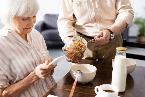 Foco seletivo da mulher idosa usando smartphone perto do marido segurando frasco de cereais em casa — Fotografia de Stock
