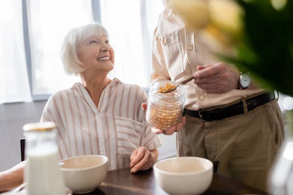 Selective focus of senior woman smiling at husband pouring cereals from jar during breakfast — Stock Photo
