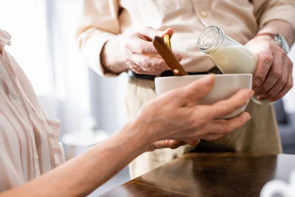 Cropped view of senior man pouring milk while wife holding bowl at table — Stock Photo