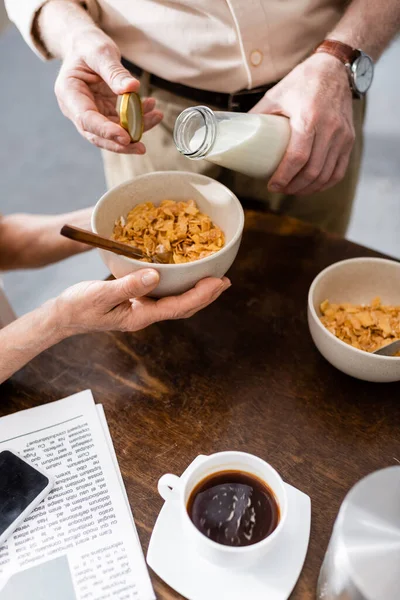 Cropped view of woman holding bowl with cereals while husband pouring milk near coffee, newspaper and smartphone on table — Stock Photo