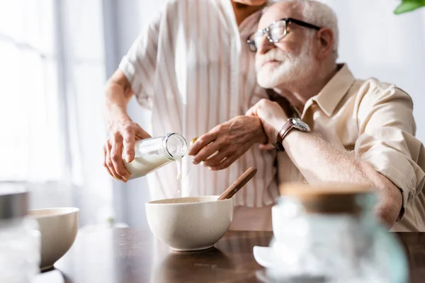 Selective focus of man hugging senor wife pouring milk in bowl at table — Stock Photo