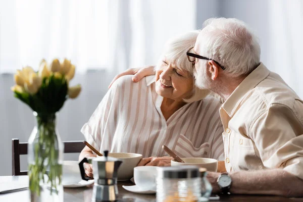 Foco seletivo do homem sênior beijando esposa sorridente perto do café da manhã na mesa da cozinha — Fotografia de Stock