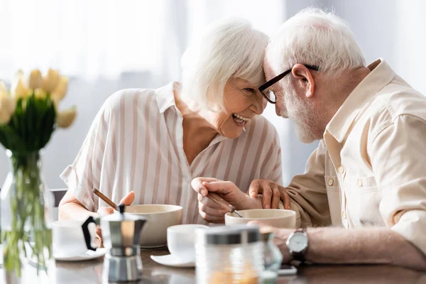 Селективный фокус позитивной пожилой пары, смотрящей друг на друга рядом с кофе и завтраком на столе — стоковое фото
