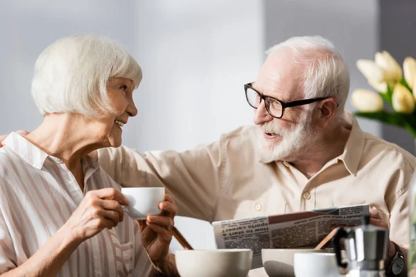 Selective focus of senior man holding newspaper and embracing smiling wife with cup of coffee — Stock Photo