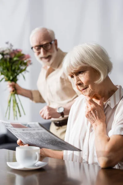 Selektiver Fokus trauriger Seniorin beim Lesen von Nachrichten neben positivem Ehemann mit Blumenstrauß zu Hause — Stockfoto