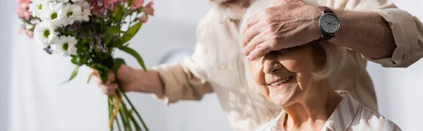 Panoramic shot of senior man covering eyes to smiling wife and holding floral bouquet at home — Stock Photo