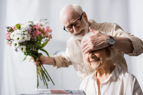 Selective focus of smiling senior man covering eyes of wife and holding bouquet — Stock Photo