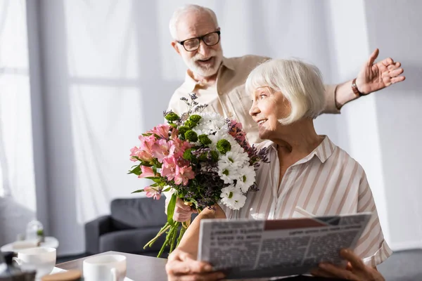 Focus selettivo dell'uomo anziano positivo che tiene il bouquet vicino alla moglie con giornale in cucina — Foto stock