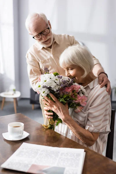 Concentration sélective de l'homme âgé embrassant femme avec bouquet près du journal et tasse de café sur la table — Photo de stock