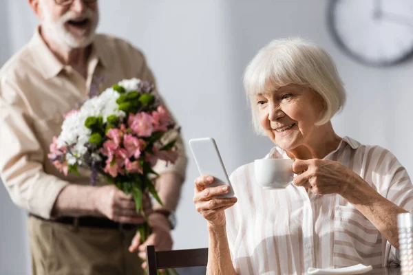 Selective focus of smiling woman holding cup of coffee and using smartphone near husband with bouquet — Stock Photo