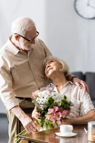 Focus selettivo dell'uomo anziano che abbraccia la moglie sorridente con bouquet vicino al caffè sul tavolo — Foto stock