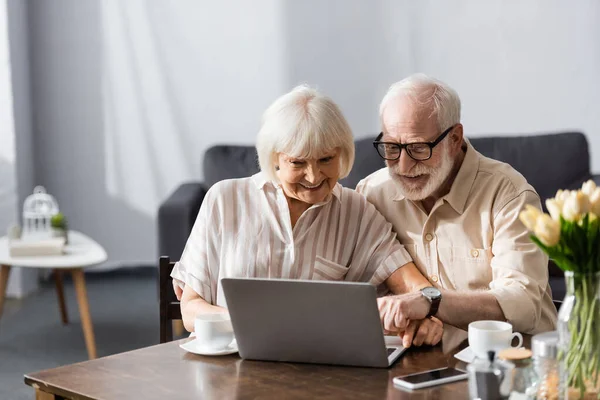 Selective focus of smiling elderly couple using laptop near coffee cups on table — Stock Photo