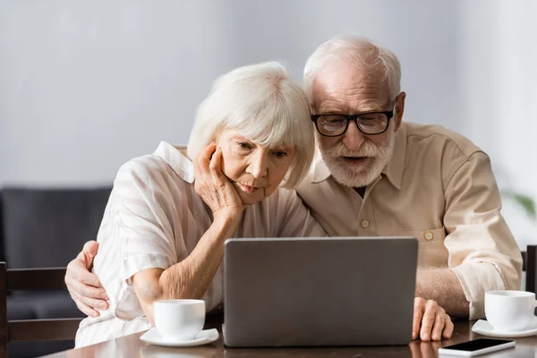 Enfoque selectivo del hombre mayor abrazando esposa mientras usa el ordenador portátil cerca de tazas de café y teléfono inteligente en la mesa - foto de stock