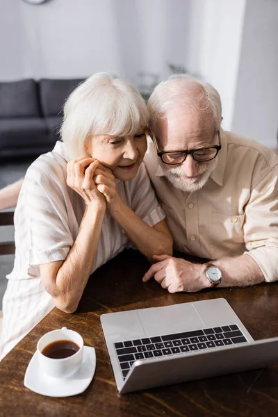 Selektiver Fokus des Seniorenpaares beim Blick auf den Laptop neben der Kaffeetasse auf dem heimischen Tisch — Stockfoto