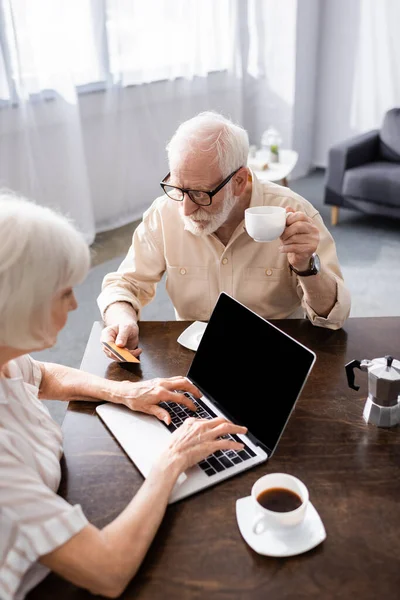 Focus selettivo dell'uomo anziano in possesso di carta di credito e tazza di caffè vicino alla moglie utilizzando il computer portatile a casa — Foto stock