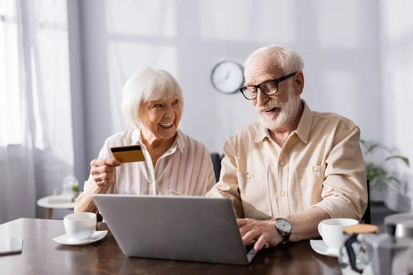 Enfoque selectivo de la pareja mayor sonriente utilizando el ordenador portátil y la tarjeta de crédito cerca de tazas de café en la mesa - foto de stock