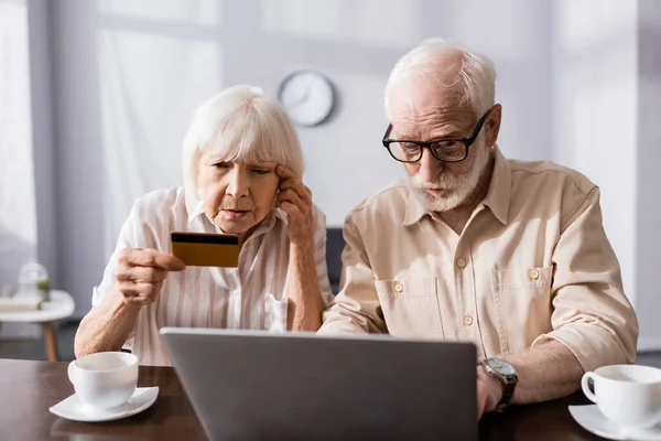 Focus selettivo della donna anziana guardando la carta di credito vicino all'uomo utilizzando laptop e tazze sul tavolo — Foto stock