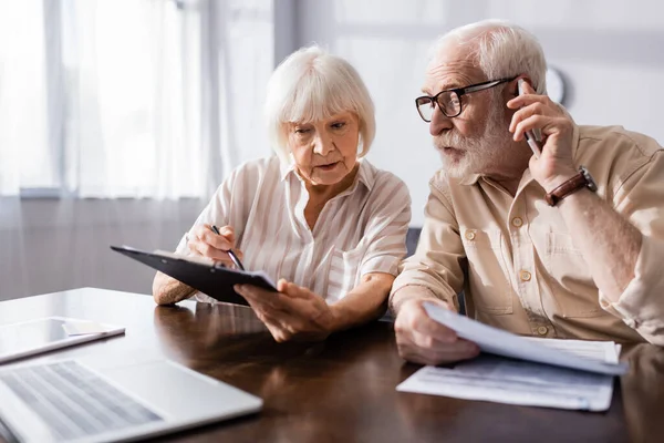 Выборочный фокус разговора пожилого человека на смартфоне рядом с женой с документами — стоковое фото
