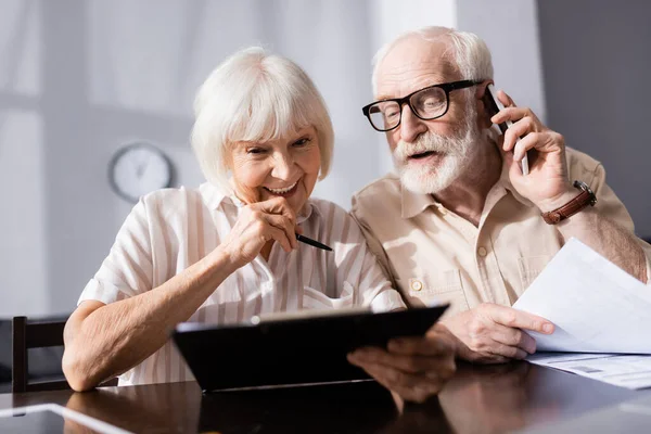 Selektiver Fokus einer lächelnden Seniorin mit Klemmbrett in der Nähe ihres Mannes, der zu Hause auf dem Smartphone spricht — Stockfoto