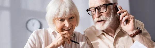 Panoramic shot of senior man talking on smartphone near wife holding pen — Stock Photo