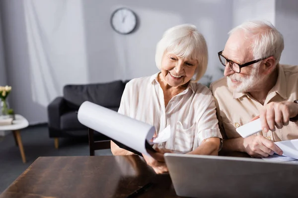 Selective focus of smiling woman holding clipboard with papers beside husband holding smartphone and laptop on table — Stock Photo