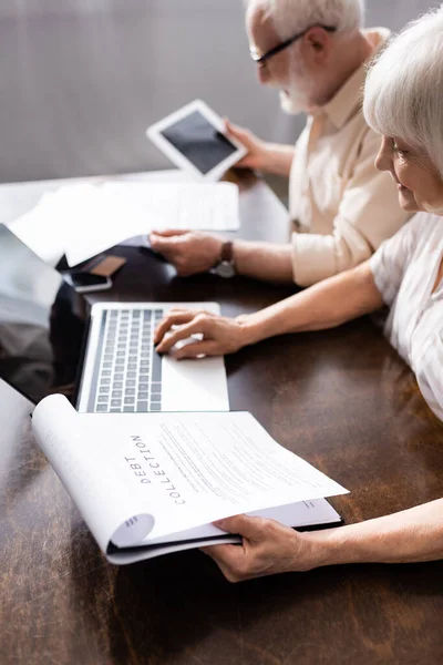 Selective focus of senior woman holding papers with debt collection lettering and using laptop near husband at home — Stock Photo