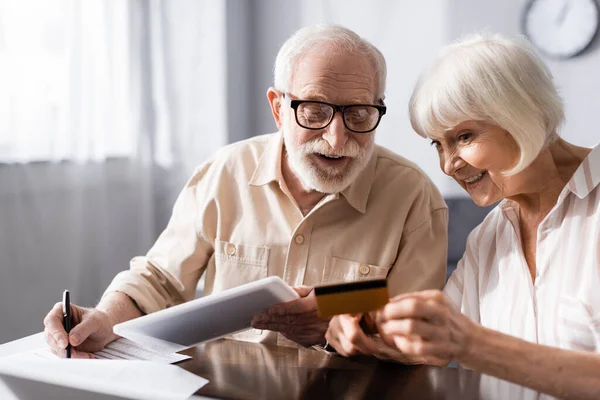 Enfoque selectivo de la mujer mayor sonriente sosteniendo la tarjeta de crédito cerca del marido con la escritura digital de la tableta en papeles - foto de stock