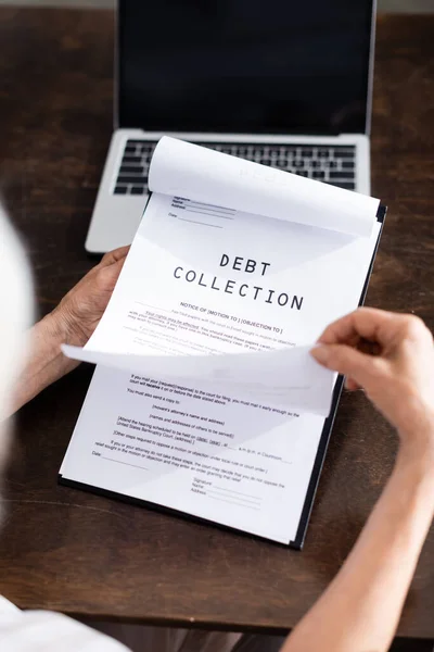 Selective focus of elderly woman holding papers with debt collection lettering near laptop on table — Stock Photo