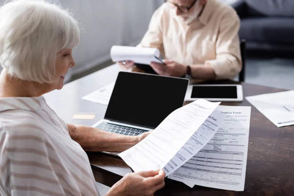 Selektiver Fokus einer lächelnden Seniorin mit Papieren in der Nähe von Geräten und Kreditkarten auf dem Tisch — Stockfoto