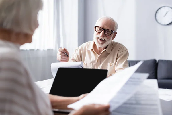 Focus selettivo dell'uomo sorridente che guarda la moglie con documenti vicino al computer portatile a casa — Foto stock