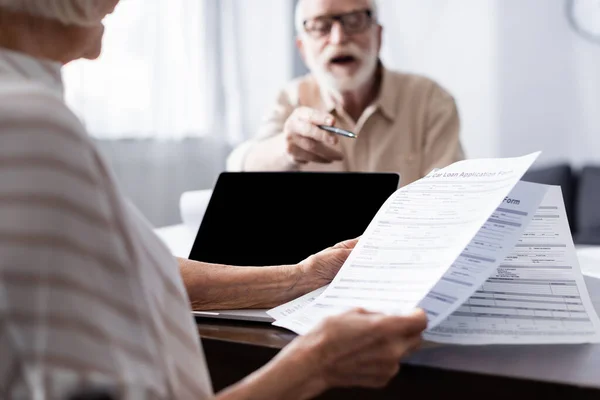 Selektiver Fokus eines älteren Mannes, der auf Frau zeigt, die Papiere in der Nähe von Laptop auf Tisch hält — Stockfoto