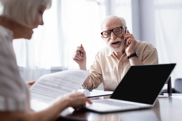Selective focus of smiling senior man talking on smartphone near wife holding papers and using laptop at home — Stock Photo