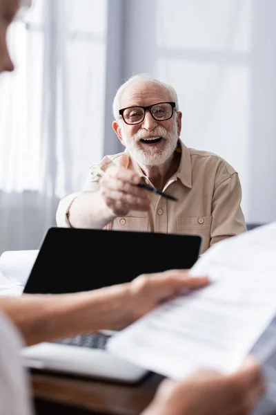 Вибірковий фокус позитивного літнього чоловіка, який тримає ручку біля дружини з паперами та ноутбуком на столі — стокове фото