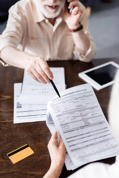 Выборочный фокус человека, говорящего на смартфоне и указывающего на бумаги возле жены и кредитной карты на столе — стоковое фото
