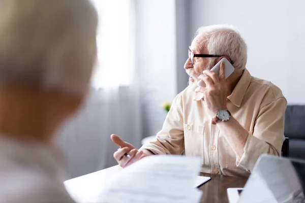 Selektiver Fokus eines älteren Mannes, der in der Nähe seiner Frau mit Smartphone und Papieren auf dem Tisch spricht — Stockfoto