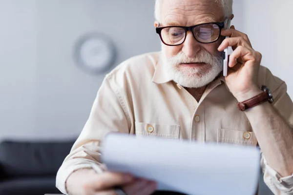 Selective focus of senior man holding document and talking on smartphone at home — Stock Photo