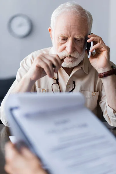 Selective focus of sad senior man talking on smartphone near woman holding papers — Stock Photo